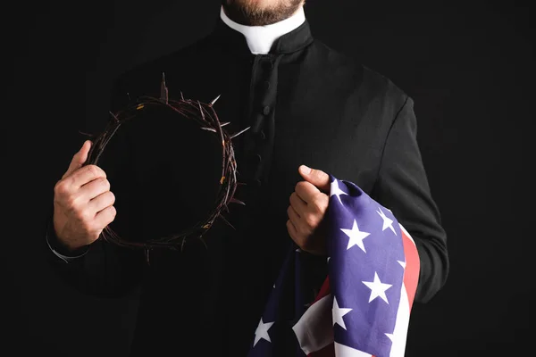 Cropped view of priest holding wreath with spikes and american flag isolated on black — Stock Photo