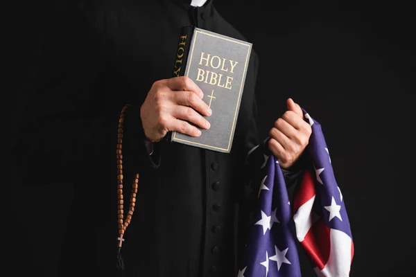 Vista recortada de sacerdote sosteniendo la sagrada Biblia, bandera americana y rosario perlas aisladas en negro - foto de stock