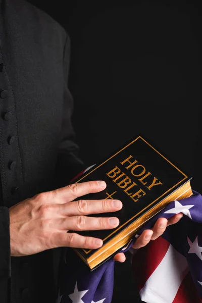Abgeschnittene Ansicht eines Priesters mit heiliger Bibel und amerikanischer Flagge isoliert auf schwarz — Stockfoto