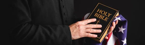 Panoramic concept of priest holding holy bible and american flag isolated on black — Stock Photo