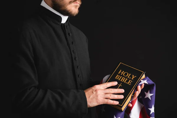 Vista parcial del sacerdote sosteniendo la sagrada Biblia y la bandera americana aislada en negro - foto de stock