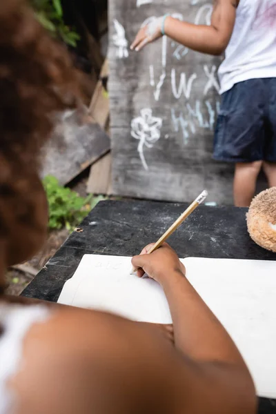Vista recortada de niño afroamericano sosteniendo lápiz mientras escribe cerca de niño y pizarra - foto de stock
