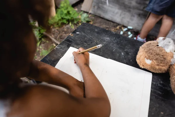 Vista parcial de un niño afroamericano sosteniendo lápiz mientras escribe cerca de un niño - foto de stock