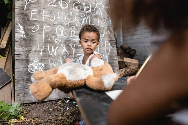 Messa a fuoco selettiva del ragazzo africano americano che tiene in mano il giocattolo morbido vicino al bambino e alla lavagna — Foto stock