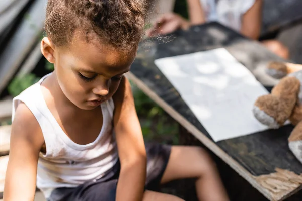 Selektiver Fokus armer afrikanischer amerikanischer Junge, der in der Nähe des Tisches mit Papier nach unten schaut — Stockfoto