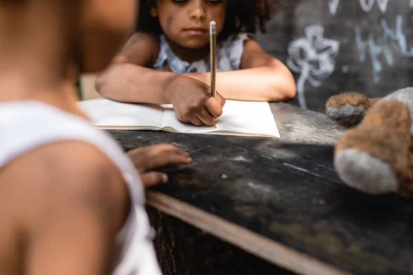 Visão cortada da criança afro-americana pobre escrevendo em papel perto do irmão — Fotografia de Stock