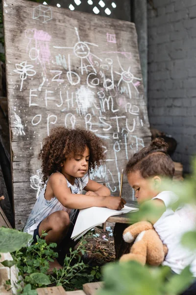 Foco seletivo de crianças afro-americanas pobres escrevendo em notebook perto de irmão e quadro-negro — Fotografia de Stock