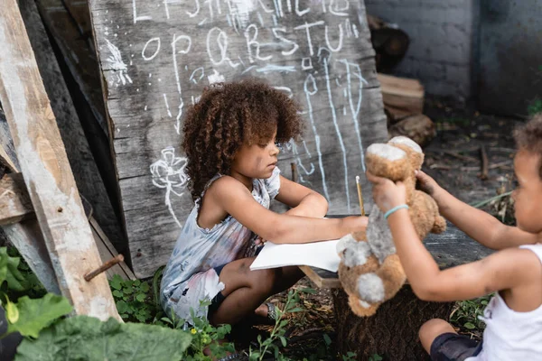 Enfoque selectivo de rizado afroamericano niño escribiendo en portátil cerca de hermano pobre jugando con juguete suave - foto de stock