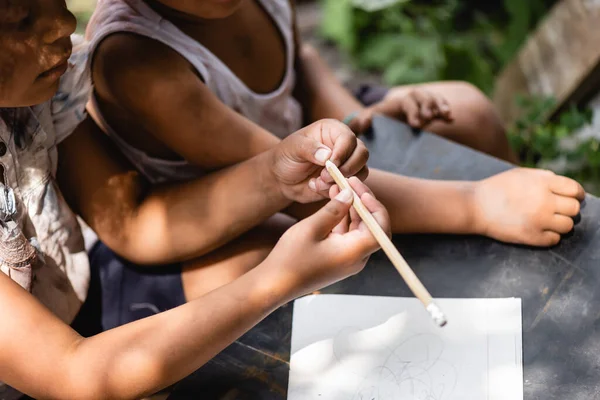 Vista recortada de un niño afroamericano pobre sosteniendo lápiz cerca de un cuaderno en el escritorio y el niño - foto de stock