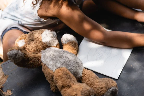 Cropped view of poor african american kid near teddy bear and notebook on desk — Stock Photo