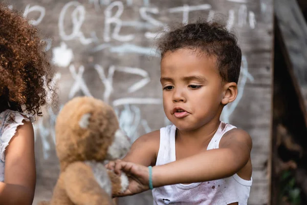 Foco seletivo do pobre menino afro-americano brincando com urso de pelúcia sujo perto da criança — Fotografia de Stock