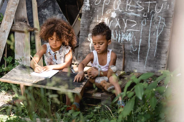 Focalizzazione selettiva del bambino africano americano che scrive vicino al fratello seduto con orsacchiotto sporco — Foto stock