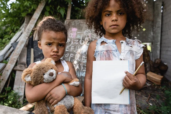 Pobre afroamericano niño sosteniendo en blanco papel y lápiz cerca molesto hermano con osito de peluche - foto de stock