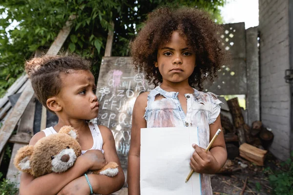 Trauriges und armes afrikanisch-amerikanisches Kind hält leeres Papier und Bleistift in der Nähe von Bruder mit Teddybär — Stockfoto