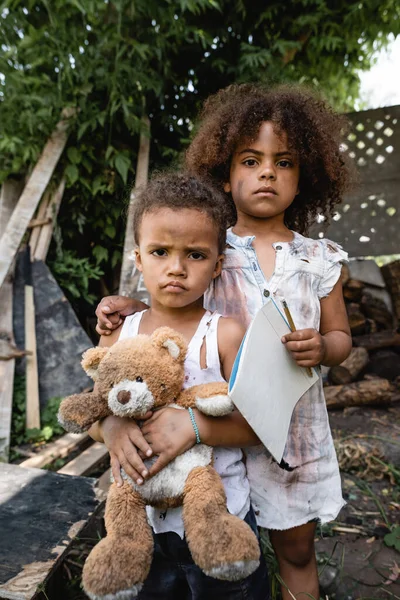 Poor african american kid holding blank paper and pencil while standing near sad brother with teddy bear — Stock Photo