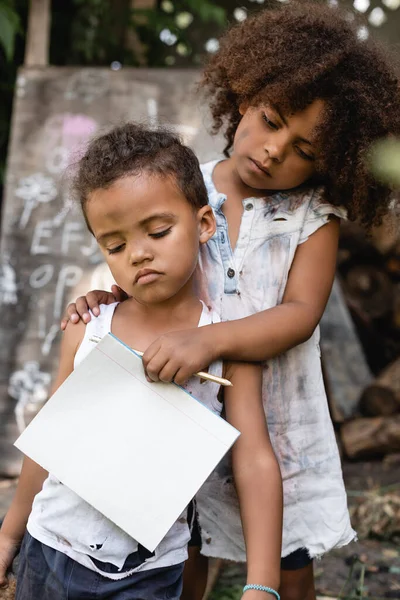 Poor african american kid holding blank notebook and pencil while touching sad brother in torn clothes — Stock Photo