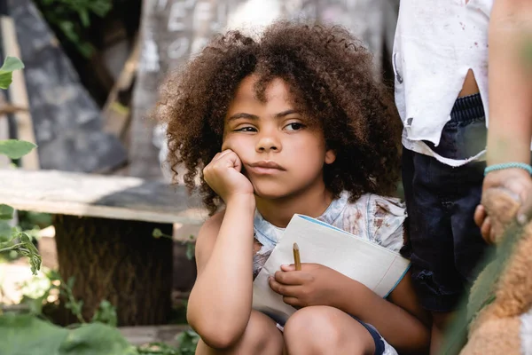 Pobre criança afro-americana segurando notebook perto de criança de pé com brinquedo macio sujo — Fotografia de Stock