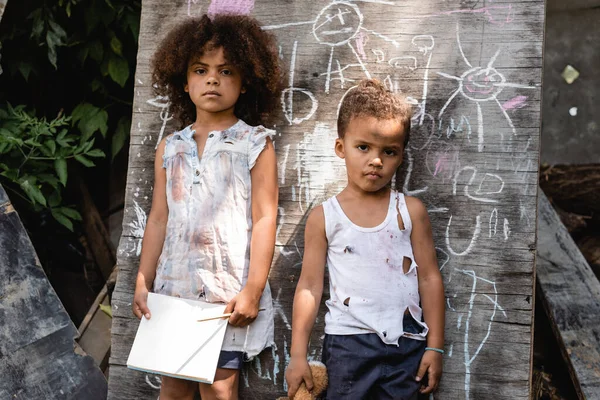 Poor african american kid in torn clothes standing with sad sister near chalkboard — Stock Photo