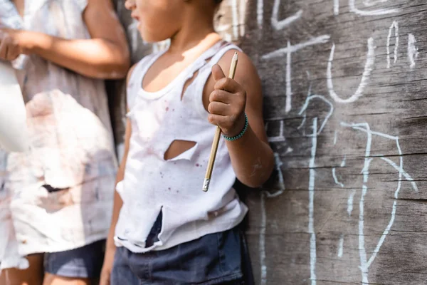 Cropped view of poor african american kid in torn clothes standing near chalkboard and sister while holding pencil — Stock Photo