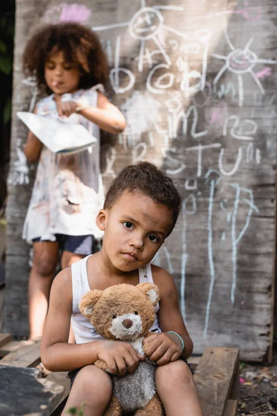Foco seletivo de criança afro-americana pobre em roupas rasgadas sentado com brinquedo macio perto de quadro-negro e irmã — Fotografia de Stock