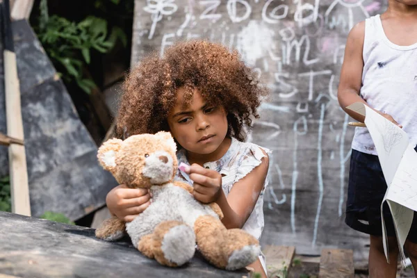 Enfoque selectivo de niño afroamericano pobre mirando oso de peluche sucio cerca de niño y pizarra - foto de stock