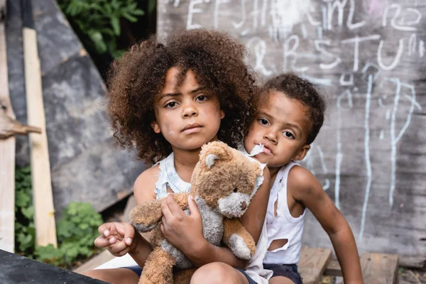 Pobres niños afroamericanos con oso de peluche sucio mirando a la cámara - foto de stock