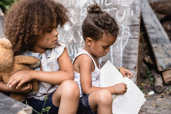 Messa a fuoco selettiva di povero ragazzo afroamericano guardando fratello in possesso di notebook e matita — Foto stock