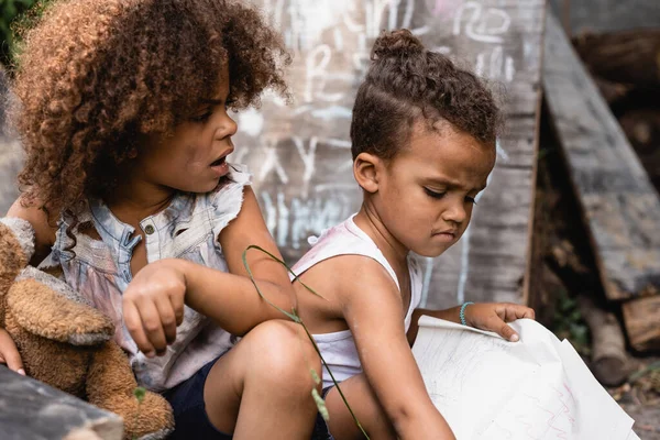 Enfoque selectivo de niño afroamericano pobre con la boca abierta mirando hermano sosteniendo papel - foto de stock