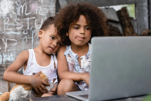 Selektiver Fokus armer afrikanisch-amerikanischer Kinder, die ihre Zunge in der Nähe ihres Bruders ausstrecken und Laptop draußen benutzen — Stockfoto