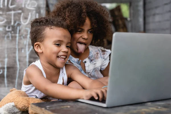 Selective focus of poor african american kid sticking out tongue near happy kid using laptop outside — Stock Photo