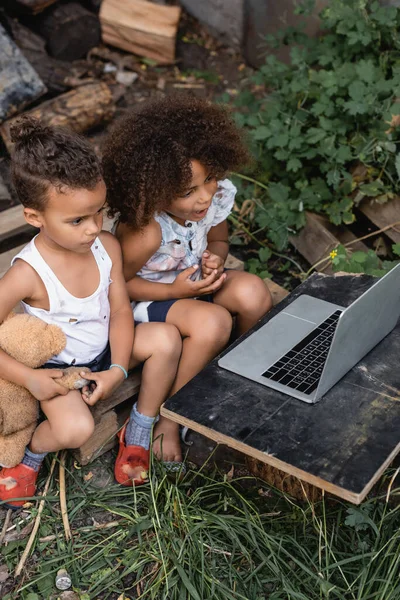 Blick aus der Vogelperspektive auf arme afrikanisch-amerikanische Kinder in zerrissenen Kleidern, die draußen auf Laptop schauen — Stockfoto