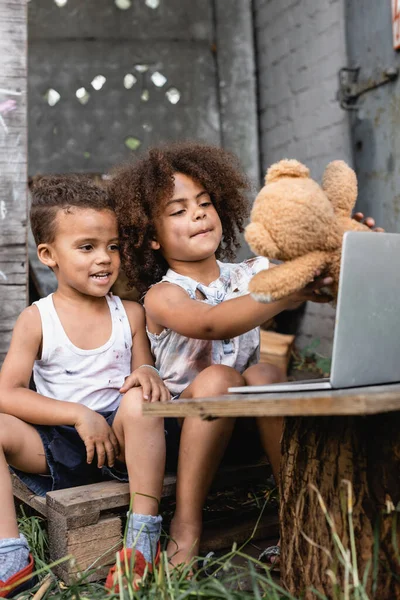 Enfoque selectivo de los niños afroamericanos pobres sosteniendo oso de peluche sucio cerca de la computadora portátil fuera - foto de stock
