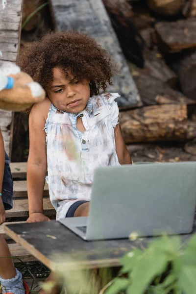 Enfoque selectivo de niño afroamericano pobre y escéptico mirando portátil cerca de hermano en la calle urbana - foto de stock