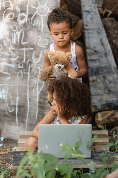 Селективный фокус грязных африканских американских детей с ноутбуком и плюшевым мишкой на городской улице — стоковое фото