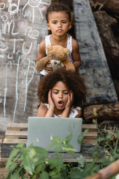 Selective focus of shocked african american child looking at laptop near brother holding teddy bear on urban street — Stock Photo