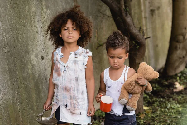 Pauvre enfant américain arican tenant cuillère en métal et assiette près de frère triste avec ours en peluche sale et tasse dans le bidonville — Photo de stock