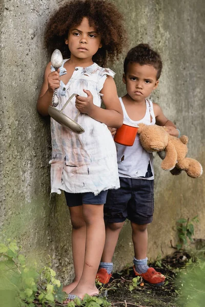 Enfoque selectivo de niños afroamericanos sin hogar sosteniendo plato y taza mientras mendigan limosnas en barrios marginales - foto de stock