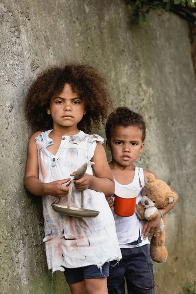 Destitute niños afroamericanos con placa y cuchara mirando a la cámara mientras mendigan limosnas cerca del muro de hormigón en la calle urbana - foto de stock