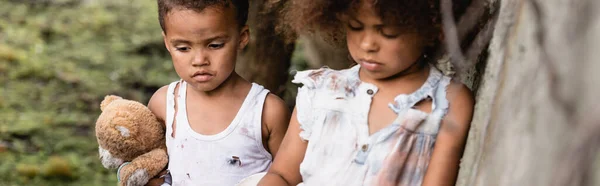 Panoramic crop of sad homeless afican american kids with dirty teddy bear standing near concrete wall in slum — Stock Photo