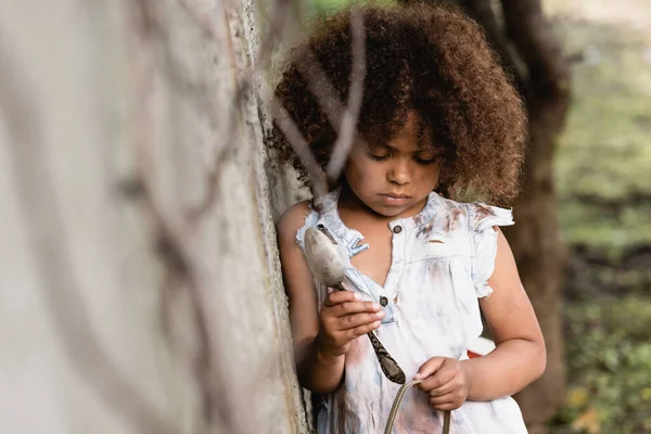 Foco seletivo de criança afro-americana pobre e chateada segurando colher de metal sujo e prato na favela — Fotografia de Stock