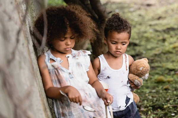 Enfoque selectivo de niños afroamericanos indefensos con cuchara, taza y oso de peluche de pie cerca de la pared de hormigón en la calle urbana - foto de stock