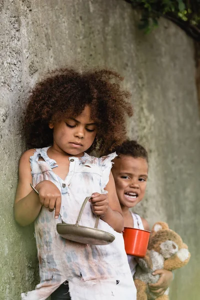 Concentration sélective d'un enfant afro-américain pauvre tenant une cuillère et une assiette en métal près d'un frère mendiant l'aumône dans un bidonville — Photo de stock