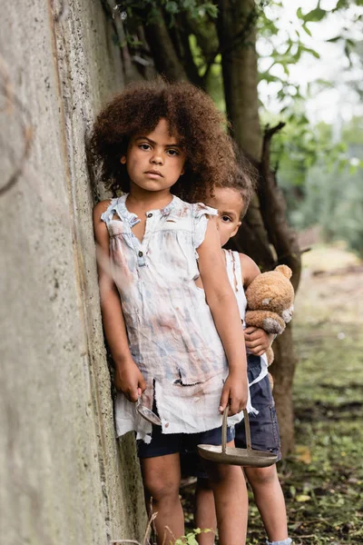 Selective focus of messy african american kids with metal plate, spoon and teddy bear looking at camera near concrete wall in slum — Stock Photo