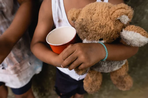 Vista recortada de niños afroamericanos sucios con oso de peluche y taza pidiendo limosna en la calle urbana - foto de stock