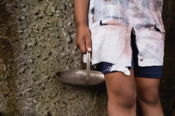 Vista recortada de un niño afroamericano pobre sosteniendo placa de metal sucio cerca de la pared de hormigón en la calle urbana - foto de stock