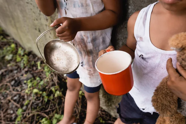 Vista ritagliata di senzatetto afro-americani bambini in possesso di tazza e piastra metallica mentre elemosina sulla strada urbana — Foto stock