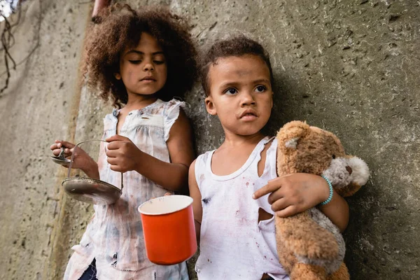 Selective focus of poor african american boy holding dirty teddy bear while begging alms near sister on urban street — Stock Photo