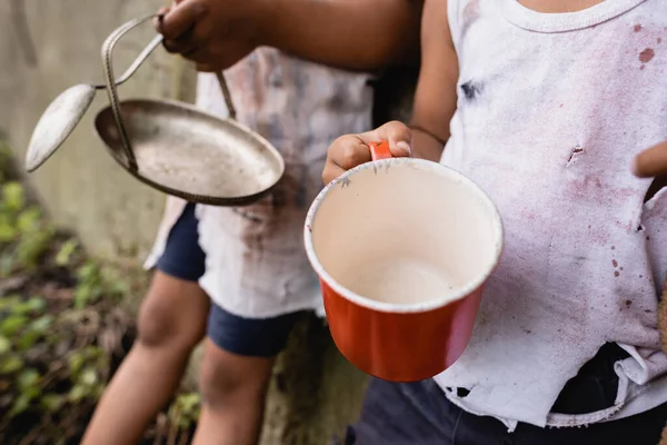 Vista recortada de niños afroamericanos pobres con taza y placa de metal de pie en la calle urbana - foto de stock