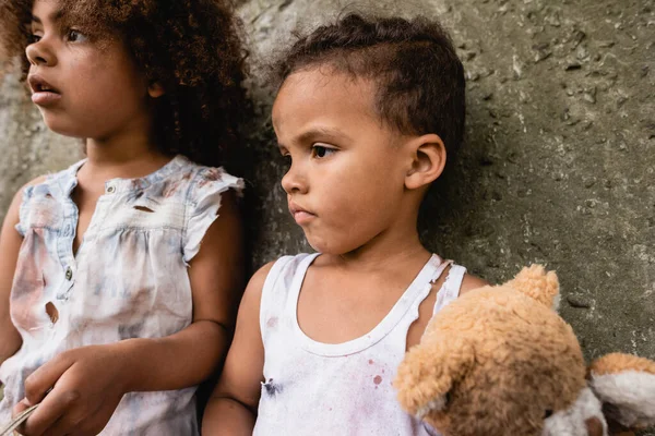 Selective focus of poor african american kids in messy clothes with teddy bear standing near concrete wall on urban street — Stock Photo