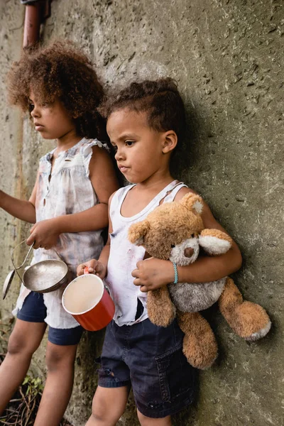 Selective focus of sad african american boy holding dirty teddy bear while begging alms near sister and concrete wall in slum — Stock Photo